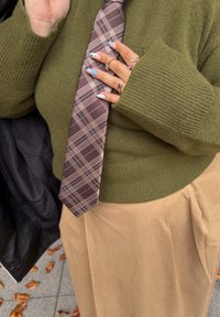 Person med en olivengrønn strikkegenser og khakifarget skjørt, som holder et brunrutete slips med velstelte negler og ringer på hånden.