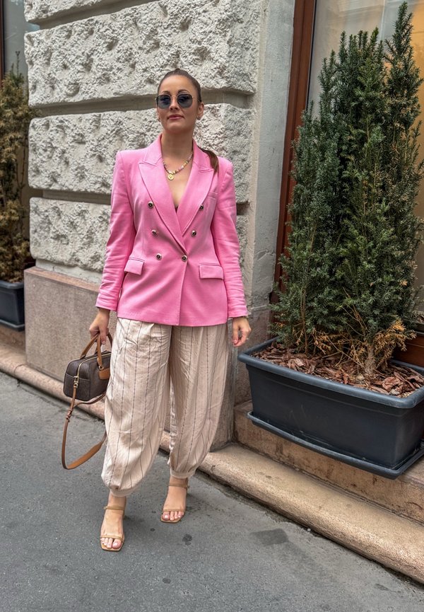 Mujer con blazer rosa brillante, pantalones beige con rayas y gafas de sol, sosteniendo un bolso marrón, de pie junto a una gran planta verde en maceta.