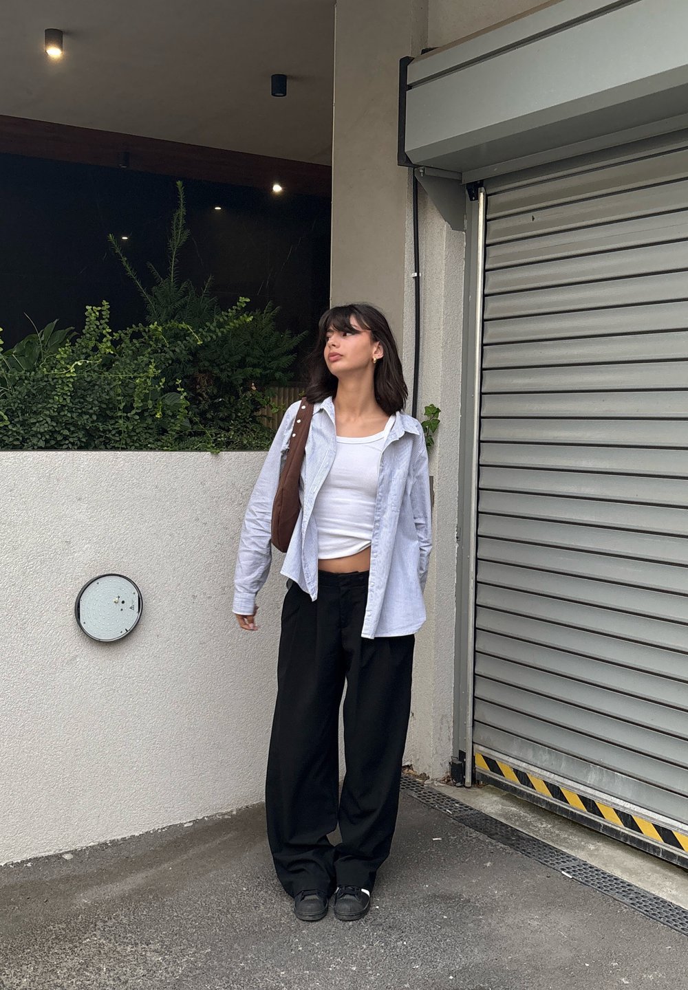 Top branco sem mangas por baixo de uma camisa de botão azul clara e calças largas pretas; bolsa de ombro castanha; em pé contra uma parede texturizada.