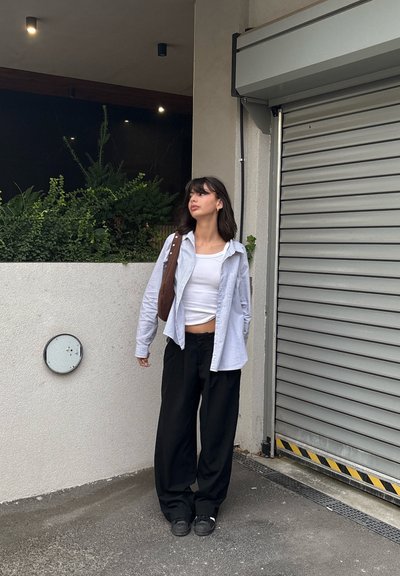 Top sin mangas blanco bajo una camisa abotonada azul clara y pantalones de pierna ancha negros; bolso marrón de hombro; de pie contra una pared texturizada.