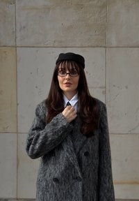 Manteau gris en laine à double boutonnage avec tissu texturé, grands boutons noirs, chemise blanche, cravate noire et béret noir. Cheveux lisses et longs.