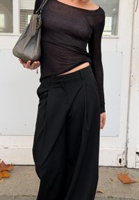 Personne portant un haut à manches longues noir transparent et un pantalon large noir, tenant un sac à main gris, debout à l'extérieur près d'une porte blanche avec des feuilles tombées à proximité.