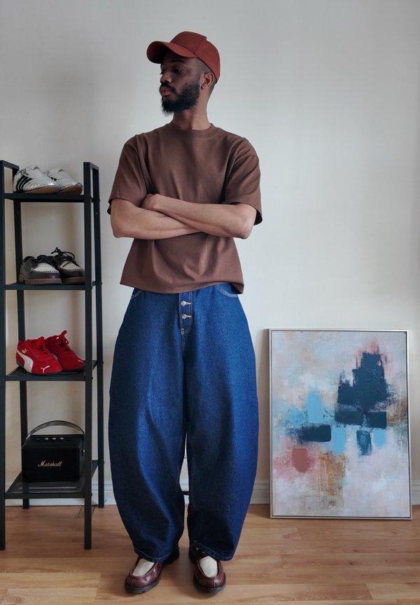 Man with beard wearing brown cap, brown shirt, wide blue jeans, standing with arms crossed near shoe rack and abstract painting.