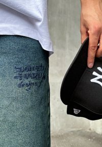 Mėlynos džinsinės kelnės su toniniais išsiuvinėtus logotipu "DON'T HARD" šalia šlaunies, derinamos su juoda kepure, laikoma rankoje.