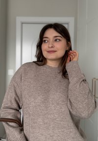 Jeune femme aux cheveux foncés portant un pull en tricot beige, debout à l'intérieur, regardant de côté avec une main touchant ses cheveux.