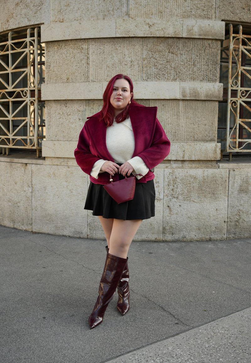 Maroon faux fur jacket over a cream sweater, black skirt, and glossy burgundy knee-high boots. Holding a small burgundy handbag. Concrete background.