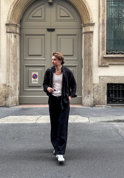 Camisa negra de botón sobre una camiseta blanca, pantalones negros y zapatillas blancas. De pie en una calle cerca de un edificio decorativo.