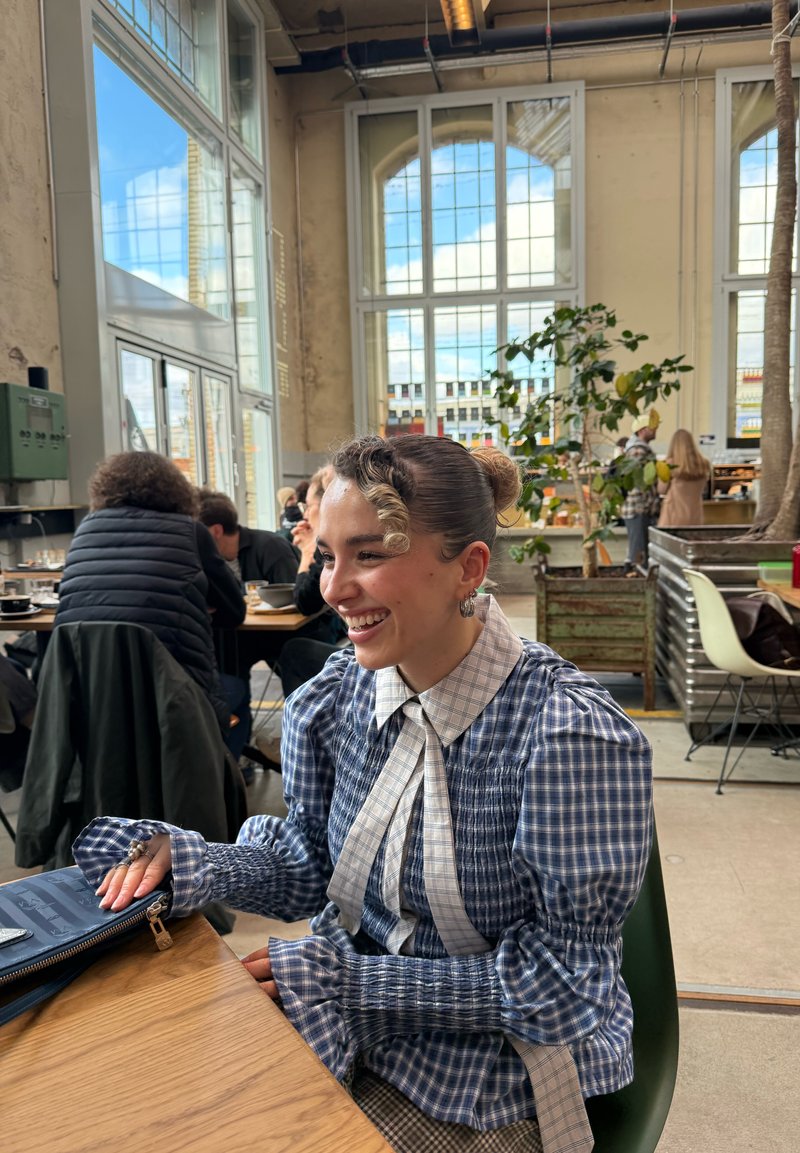 Junge Frau in einem blau-karierten Kleid, die lächelt, während sie an einem Cafétisch sitzt, mit großen Fenstern und Pflanzen im Hintergrund.