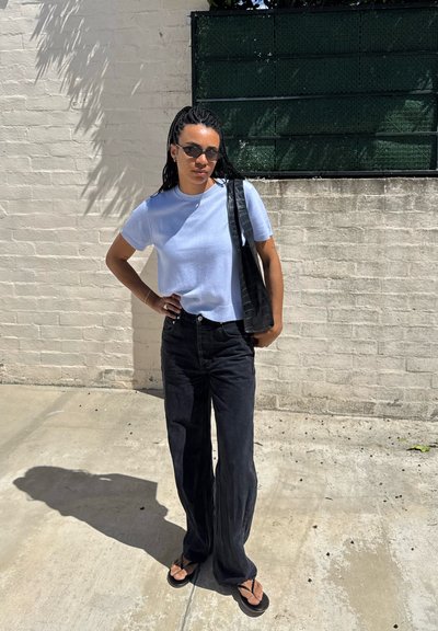 Mujer con el pelo trenzado, gafas de sol, camisa azul claro, pantalones anchos negros, chanclas y un bolso negro al hombro, de pie contra una pared de ladrillos.