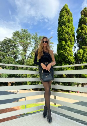 Top negro de un solo hombro combinado con una falda mini de cuero, pantys negros transparentes, tacones puntiagudos y un pequeño bolso negro de cadena.