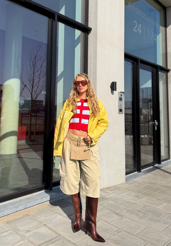 Tânără purtând o jachetă galbenă, top scurt în dungi roșii și albe, pantaloni scurți largi bej, cizme maro până la genunchi și ochelari de soare, stând în fața unei clădiri de sticlă.