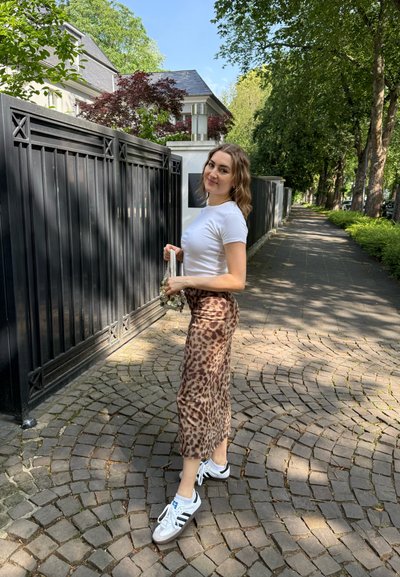 Falda larga de estampado de leopardo con un diseño fluido, combinada con una camiseta blanca ceñida y zapatillas deportivas, de pie sobre un camino de adoquines.