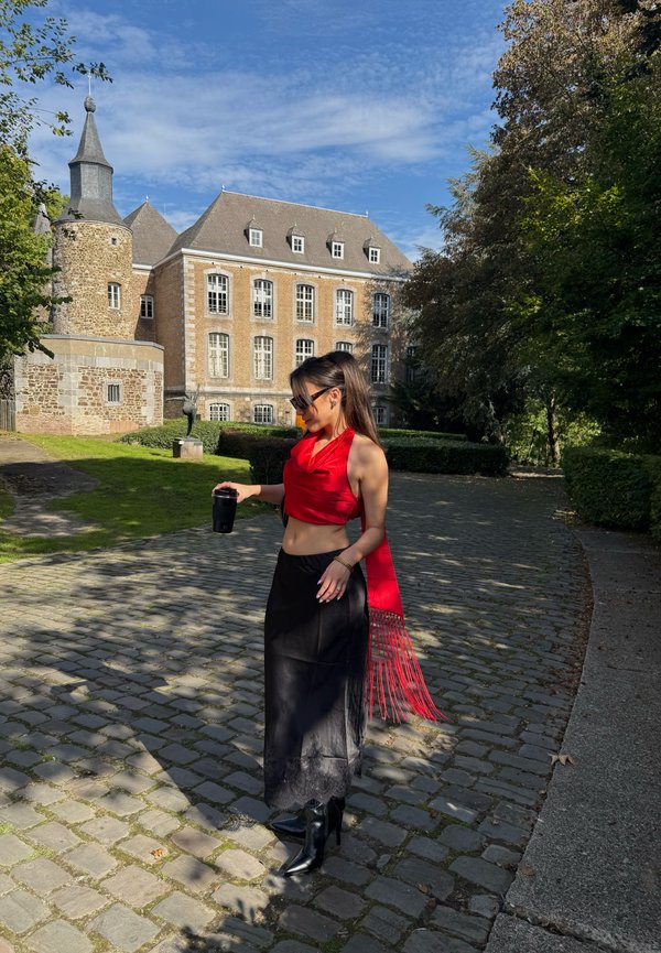 Mujer con top rojo y falda negra sosteniendo una bebida, de pie en un camino empedrado cerca de un edificio histórico de piedra bajo un cielo azul.