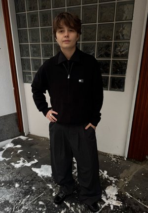 Suéter negro con cremallera sobre una camisa con cuello, combinado con pantalones oscuros plisados y zapatos negros. Presenta un pequeño logo en el suéter.