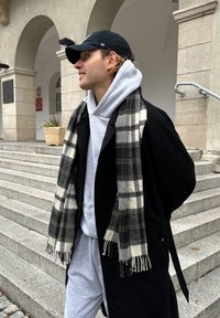 Čierny kabát s veľkými klopami cez šedú mikinu, doplnený čierno-bielym károvaným šálom. Svetlošedé teplákové nohavice a čierna čiapka. Schody v pozadí.