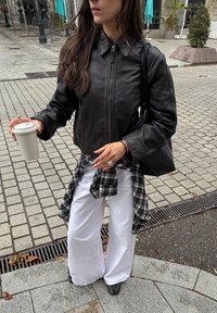 Sort læder cropped jakke med spidse krave, parret med en sort og hvid plaidskjorte knyttet om livet, og brede hvide bukser. Holder en kaffekop.