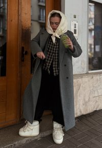 Szürke gyapjúkabát kockás inggel, fekete nadrággal és fehér platform cipővel. Zöld italt tart áttetsző pohárban.