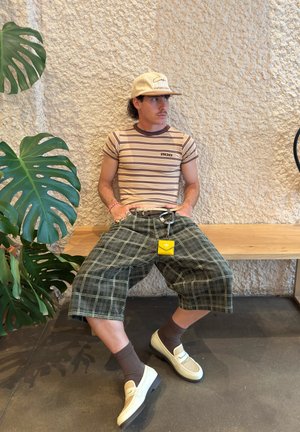 Camiseta de rayas marrón claro, pantalones cortos de cuadros oscuros, zapatos beige con malla, calcetines marrones, llavero amarillo, sentado en un banco de madera con pared texturizada.