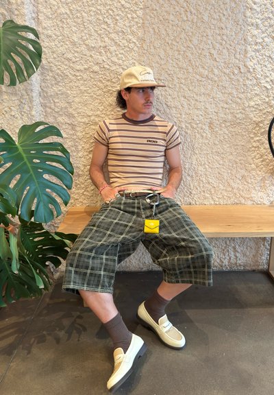 Camiseta de rayas marrón claro, pantalones cortos de cuadros oscuros, zapatos beige con malla, calcetines marrones, llavero amarillo, sentado en un banco de madera con pared texturizada.