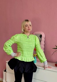 Neon zöld, ráncolt, hosszú ujjú felső fodros szegéllyel, fekete puffos szoknyával párosítva. Szürke rétegelt ujjakkal és szív medál nyaklánccal.