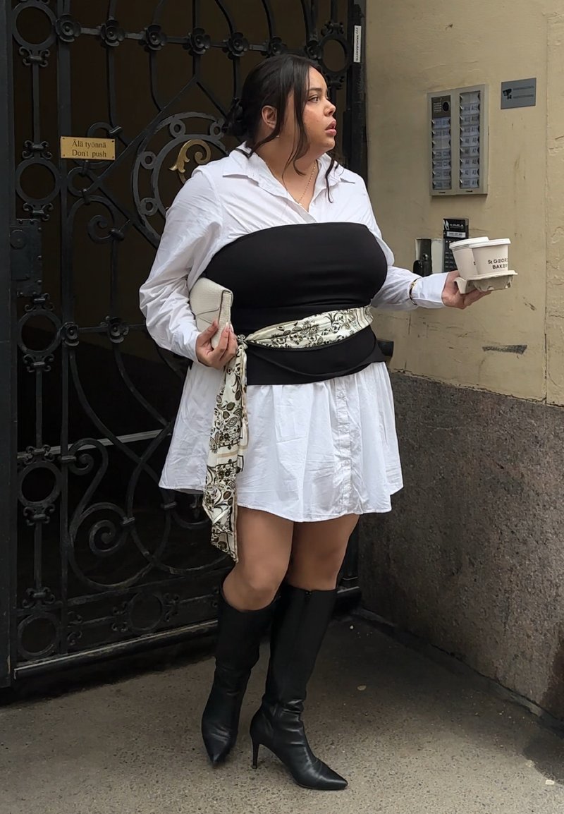 Femme en robe blanche avec un corset noir et des bottes tenant des tasses de café, debout près d'un portail en fer noir et d'un mur beige.