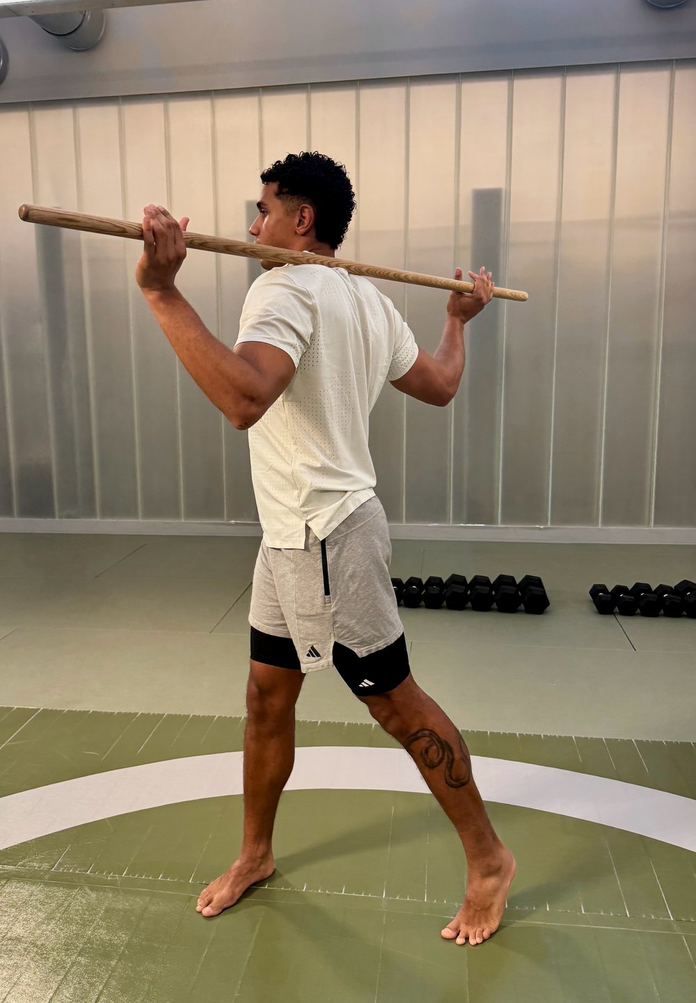 Homme en tenue de sport tenant un bâton en bois posé sur les épaules, debout pieds nus sur un tapis de gym à proximité d'haltères.