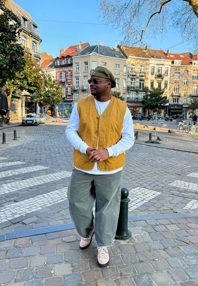 Hombre con chaleco amarillo, camisa blanca de manga larga, sombrero verde, gafas de sol y pantalones grises de pie en una calle empedrada de una ciudad europea.
