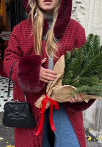 Manteau pied-de-poule rouge et noir avec bordure en fausse fourrure bordeaux, pull gris, tenant un bouquet de pin enveloppé dans du papier brun avec un ruban rouge.