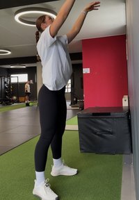 Femme en tenue de sport tendant le bras vers le haut près d'une boîte rembourrée dans une salle de sport avec un sol en gazon artificiel et du matériel d'exercice.