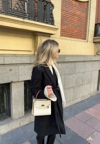 Μαύρο παλτό με ζώνη, κρεμ σάλι και κομψή τσάντα χεριού. Η τσάντα είναι λευκή με καφέ χερούλι και χρυσές λεπτομέρειες. Ο τοίχος στο φόντο είναι υφασμένος.