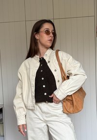 Kréma krāsas džinsu jaka ar pārmērīgu dizainu, melns teksturēts tops ar pogām, krēma krāsas bikses un brūns ādas plecu soma. Brūnas saulesbrilles.