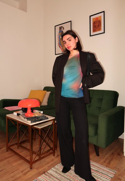 Blazer negro de gran tamaño sobre una camiseta de manga corta con degradado multicolor. Pantalones negros acampanados, de pie frente a un sofá verde y una lámpara naranja.