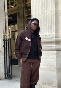 Brun zip-up hoodie med pink grafisk stjerne, parret med matchende løstsiddende brune bukser. Sort t-shirt under, solbriller, metal kædetrøje.