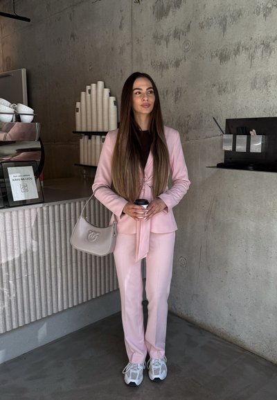 Mujer con traje rosa sosteniendo una taza de café, de pie en una cafetería moderna con vasos de papel apilados y paredes de hormigón.