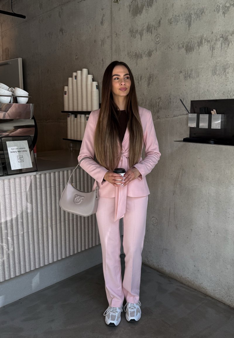 Femme en costume rose tenant une tasse de café, debout dans un café moderne avec des gobelets en papier empilés et des murs en béton.