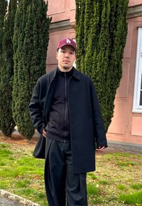 Črn plašč čez temen pulover, kombiniran z ohlapnimi črnimi hlačami. Obleko dopolnjuje bordo kapa z belim logotipom. V ozadju so zeleni grmi.