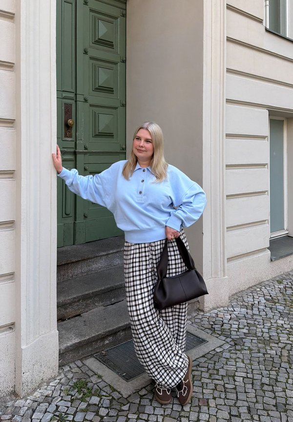Femme en pull bleu clair et pantalon à carreaux appuyée sur l'encadrement d'une porte verte dans une rue pavée, tenant un sac à main noir.