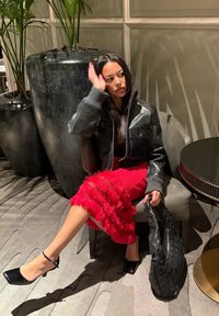 Casaco de couro preto com gola alta, saia vermelha com franjas, saltos pretos com bico quadrado e mala preta trançada. Sentada numa cadeira cinza.