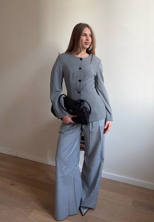 Mujer de pie en interiores, vistiendo un traje gris a medida con pantalones de pierna ancha, sosteniendo un bolso negro, mirando ligeramente de lado.