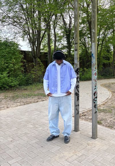 Camisa de manga corta a rayas azul claro sobre una camisa de manga larga blanca, combinada con jeans sueltos azul claro y zapatos negros. Auriculares negros puestos.