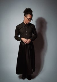 Chemise noire à manches longues avec un col pointu et des boutons, associée à une jupe fluide en satin noir. La personne porte des chaussures noires et des lunettes.