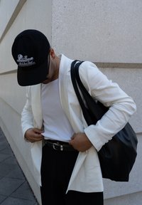 Blazer blanco sobre una camisa blanca, cinturón negro y pantalones negros. Gorra negra con un diseño gráfico. Gran bolso negro de hombro.