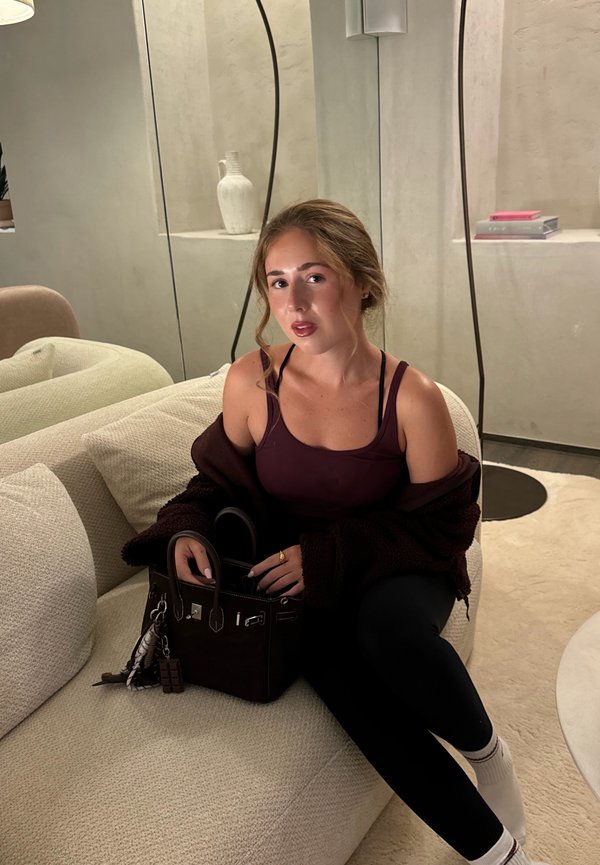 En burgunder-farvet tanktop lagd over en mørk cardigan, sorte leggings, og holder en mørk håndtaske med detaljeret hardware og en charme.