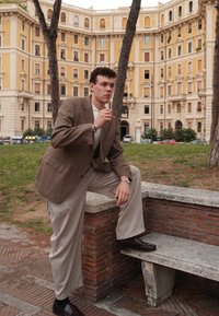 Blazer castanho com padrão, calças bege claras, sapatos escuros, em pé ao lado de um banco de pedra com relva verde e edifícios históricos ao fundo.