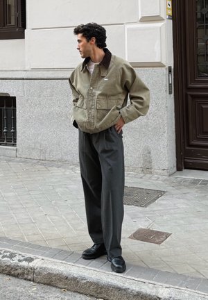 Chaqueta verde oliva con cuello marrón, dos bolsillos delanteros, combinada con pantalones plisados gris oscuro y zapatos negros, en una acera de la ciudad.