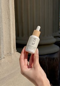 Bouteille de sérum de soin de la peau blanche avec un capuchon beige clair, présentant un texte noir. L'applicateur à goutte est visible. Une main tient la bouteille contre une surface texturée.