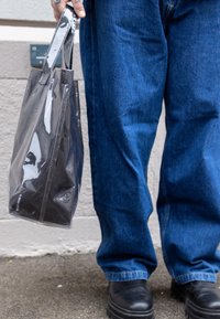 Transparentná taška na rameno z lesklého plastu s čiernymi detailmi, pevnými rukoväťami a obdĺžnikovým tvarom. Nosená s modrými džínsami a čiernymi topánkami.