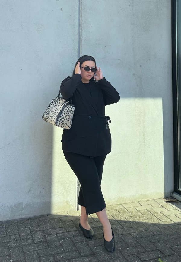 Una mujer vestida de negro se ajusta las gafas de sol junto a un muro de hormigón y lleva una bolsa de tela estampada.