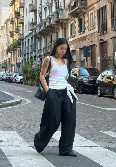 Pantalones de pierna ancha negros con un cinturón de tela blanca, combinados con una camiseta sin mangas blanca ajustada. Un bolso negro, cabello negro lacio largo y tatuajes visibles.