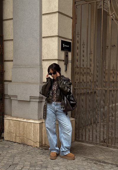 Chaqueta de cuero negra recortada, camisa de estampado de leopardo, jeans holgados azul claro, zapatos de plataforma marrones y gafas de sol negras. Fondo de pared de piedra.
